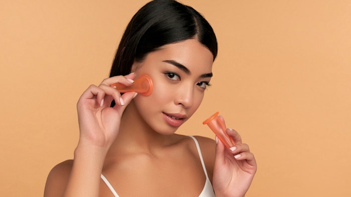 A young Asian woman with clean radiant skin makes a vacuum massage of the face with silicone facial cups on a beige background. Spa care, cupping.